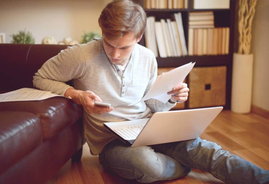 A young man is sitting in a living room with his laptop, phone and some paperwork