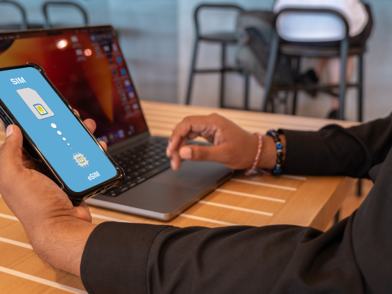 A man in business attire works at his laptop. He is holding a phone that has an eSIM installed.